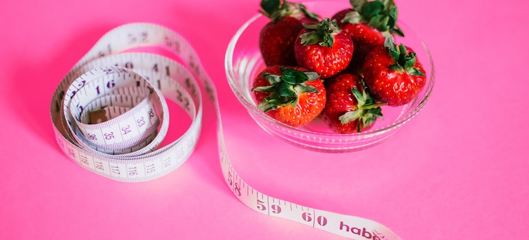 Strawberries in a bowl and a measuring tape next to it on a pink surface representing the role of nutrition in achieving your fitness goals