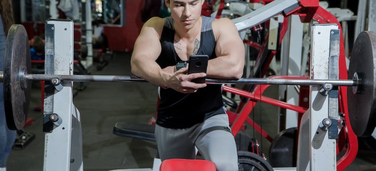 A man looking at his phone while resting on weight bar of a bench press in a gym