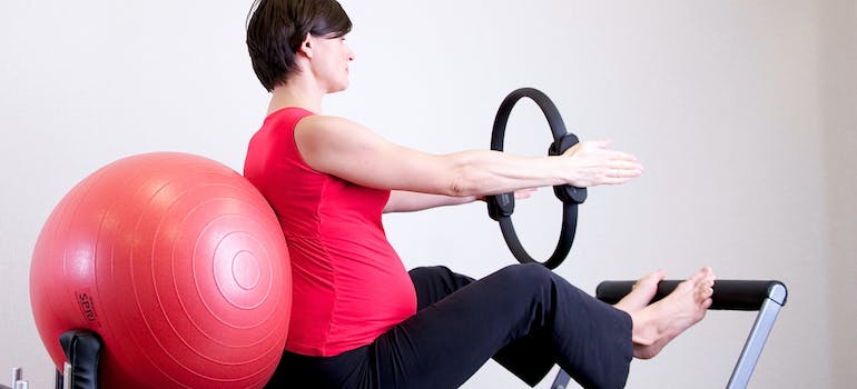 A pregnant woman exercising on a piece of fitness equipment with a prenatal personal trainer UAE
