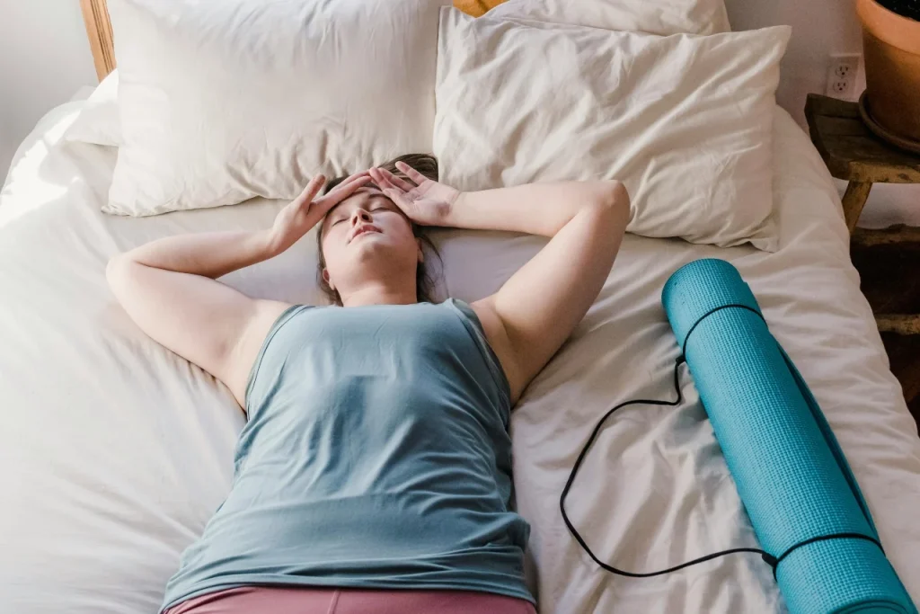 Woman lying awake on a bed with her hands on her forehead, next to a blue yoga mat and earphones.